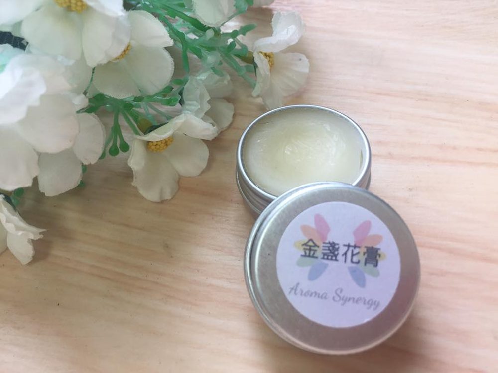 Open tin of golden calendula balm with its floral Aroma Synergy label beside white blossoms