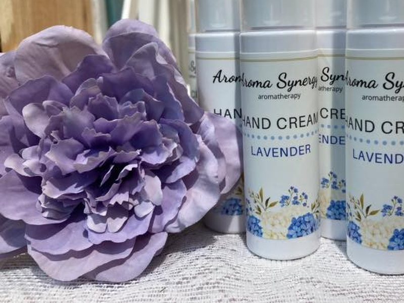 Rows of Aroma Synergy lavender hand cream bottles beside a pressed purple flower