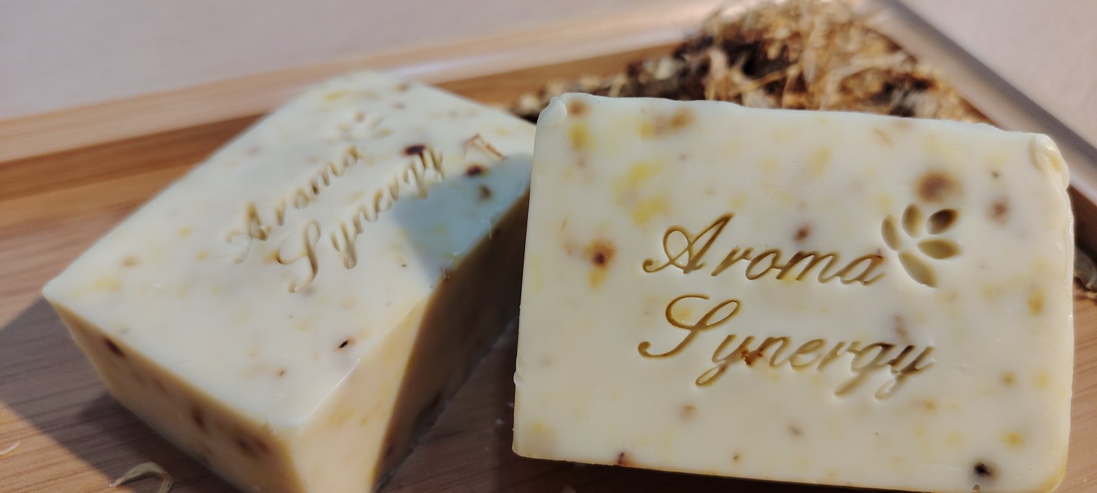 Two handmade botanical soap bars stamped with the Aroma Synergy logo on a wooden tray
